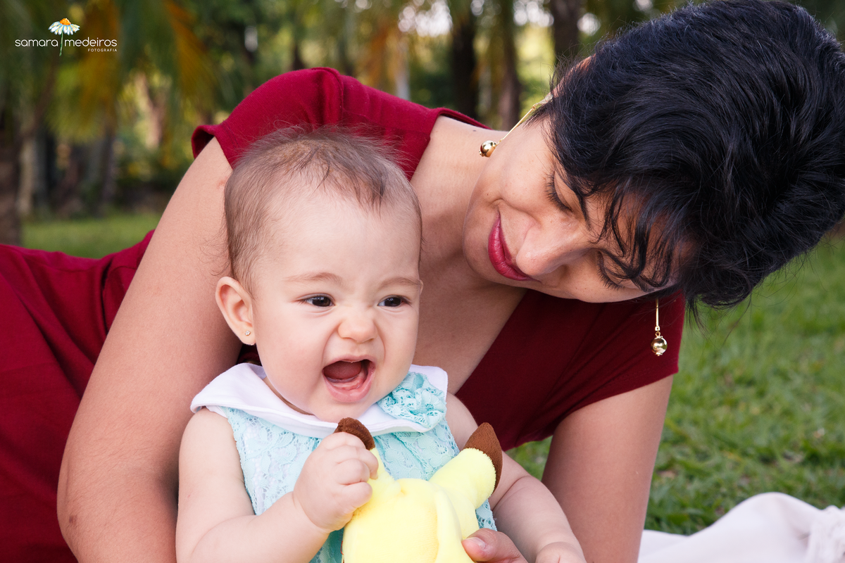 Mãe e bebê sentadas na grama brincando, a bebê sorrindo com a boca aberta e segurando um Pokemon Pikachu de pelúcia.