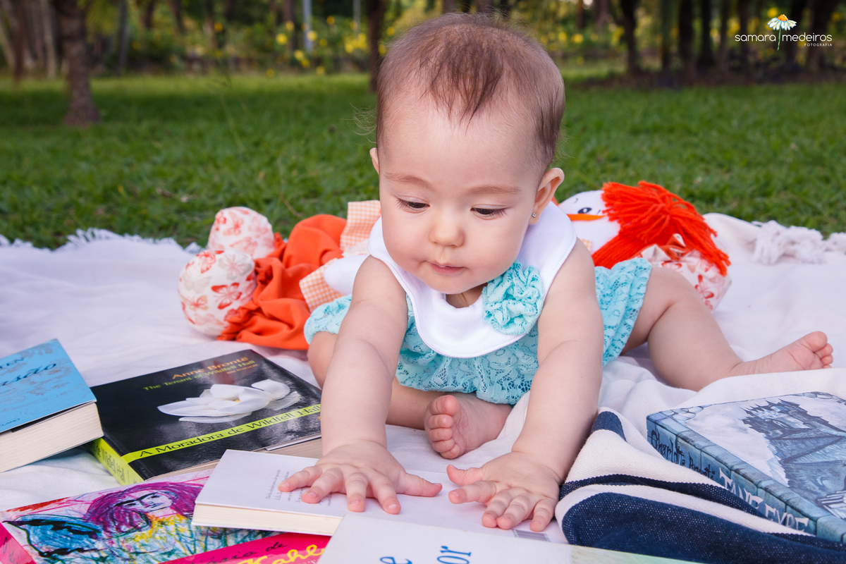Bebê sentada em uma coberta inclinada para frente olhando alguns livros de sua mãe espalhados à sua volta.