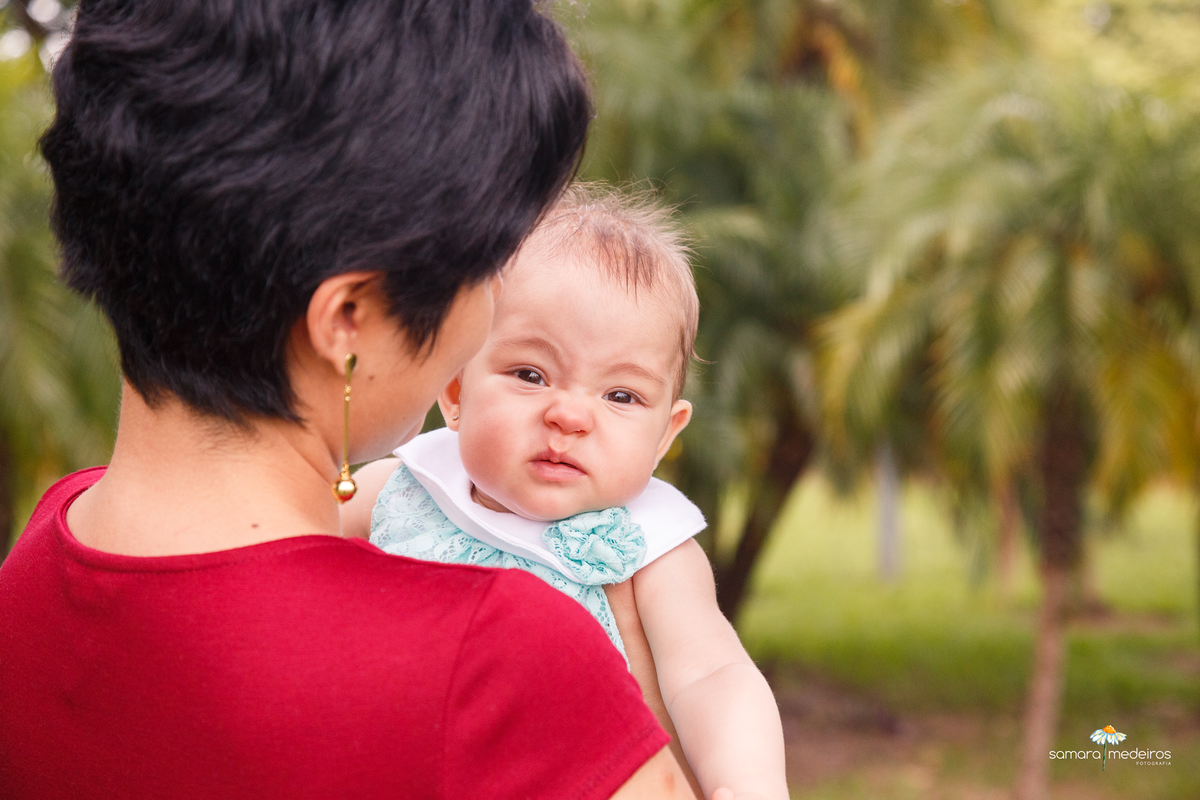 Mãe de costas para a foto e a bebê olhando para a câmera fazendo careta de bichinho, com árvores ao fundo, um pouco desfocadas.