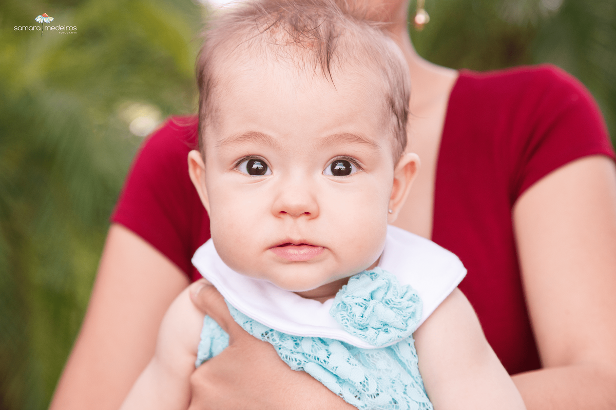 Close no rosto da criança, que olha diretamente para a câmera, no colo da mãe, que aparece apenas o tronco e braço na foto.