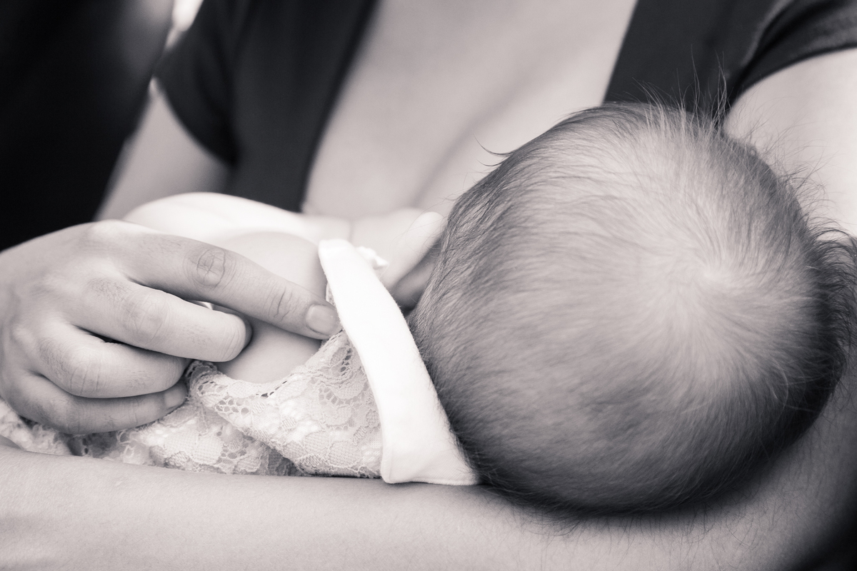Foto em preto e branco da bebê de 9 meses mamando, com foco maior na cabeça da bebê e mão da mãe fazendo carinho no braço dela.