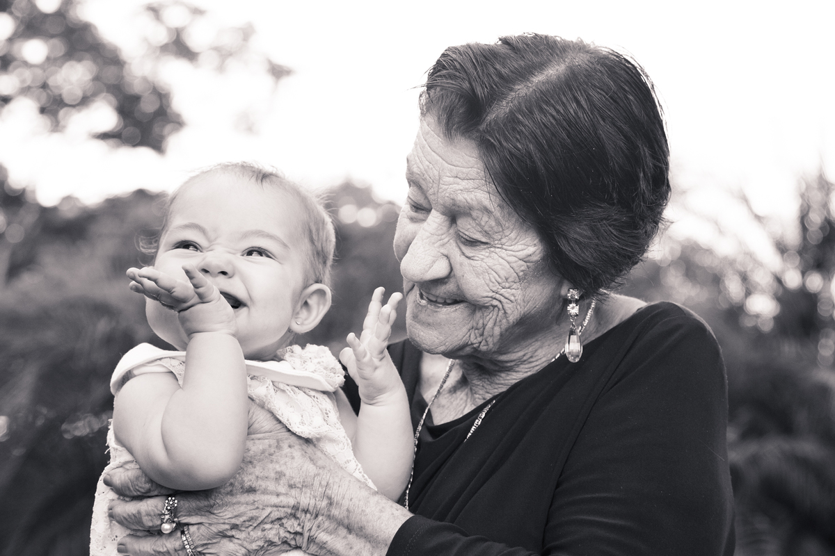Foto em preto e branco da bebê no colo da bisavó, rindo, olhando para a mãe, que não está na foto, com um dedinho na boca e sorrindo, a bisavó a observa.
