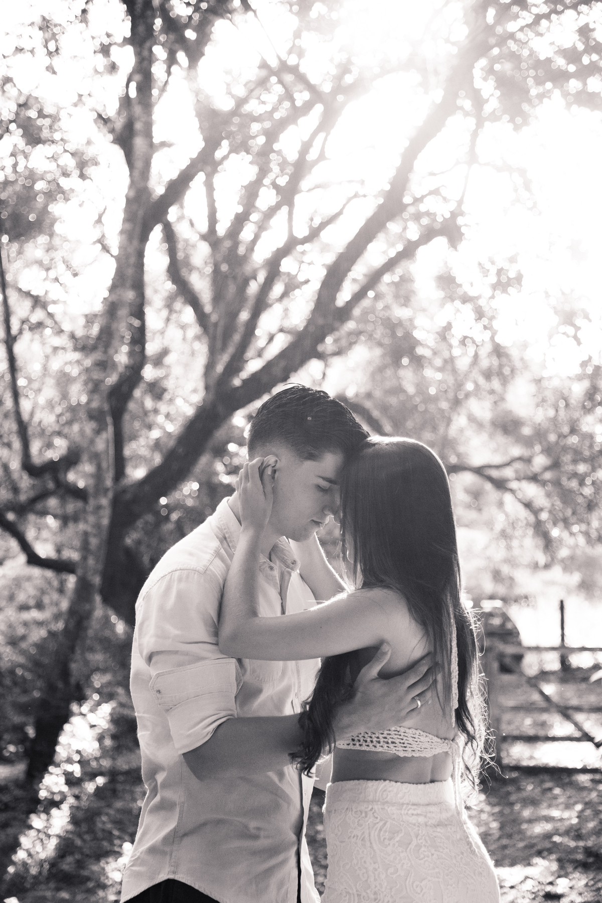 Foto em preto e branco de um casal de pé, com as testas se encontrando, com árvores ao fundo.