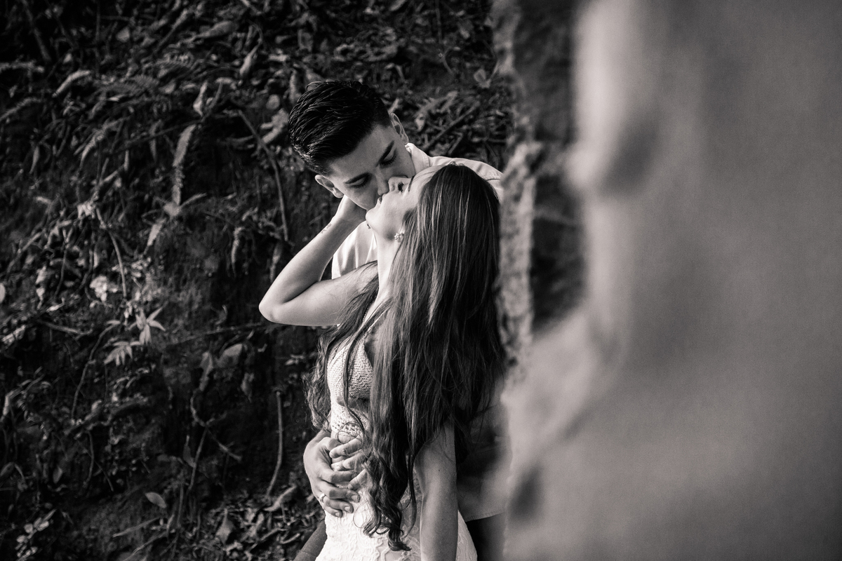 Foto em preto e branco de um casal encostado em uma parede de pedras, prestes a se beijar e a moça está com a mão levantada, apoiada no rosto do rapaz.
