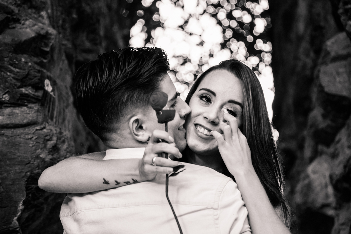 Foto em preto e branco do abraço de um casal após o pedido de casamento, o rapaz de costas, com o rosto virado de lado, para a moça e ela sorrindo e enxugando uma lágrima.
