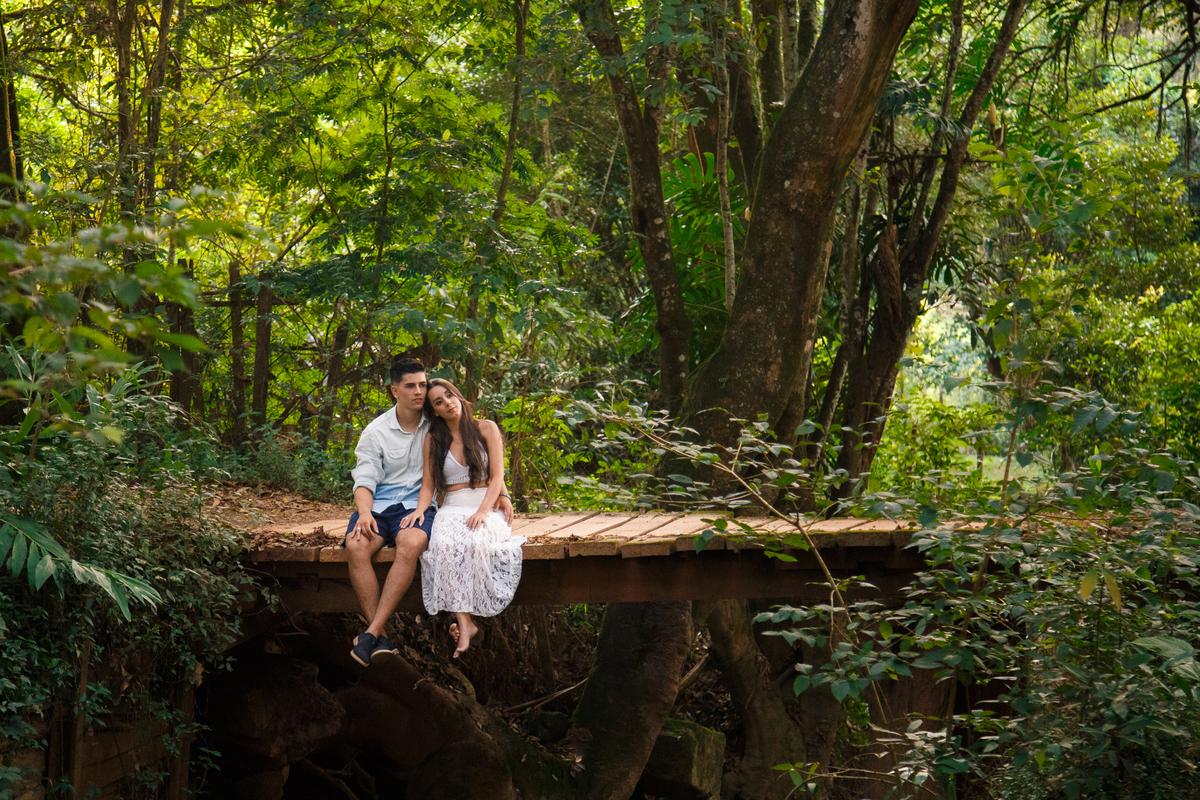 Foto aberta de um casal sentado sobre uma ponte de madeira por cima de um rio, com as pernas balançando, abraçados, observando o ambiente.