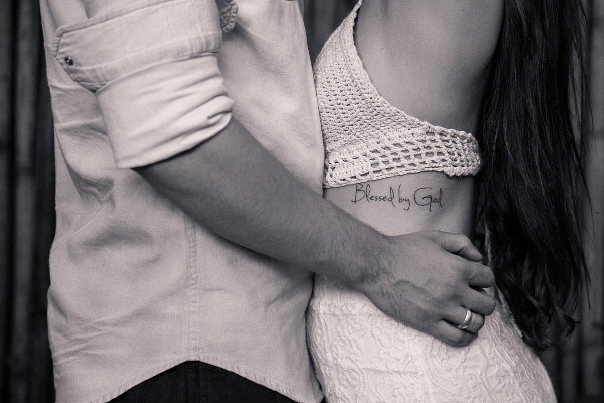 Foto em preto e branco do tronco da lateral do tronco de um casal se abraçando, o rapaz com as mãos na cintura da moça, abaixo do escrito na costela dela, Blessed by God.