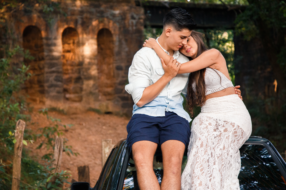 Casal se abraçando, sentados no teto do carro deles, com as ruínas de um trilho de trem abandonado ao fundo.
