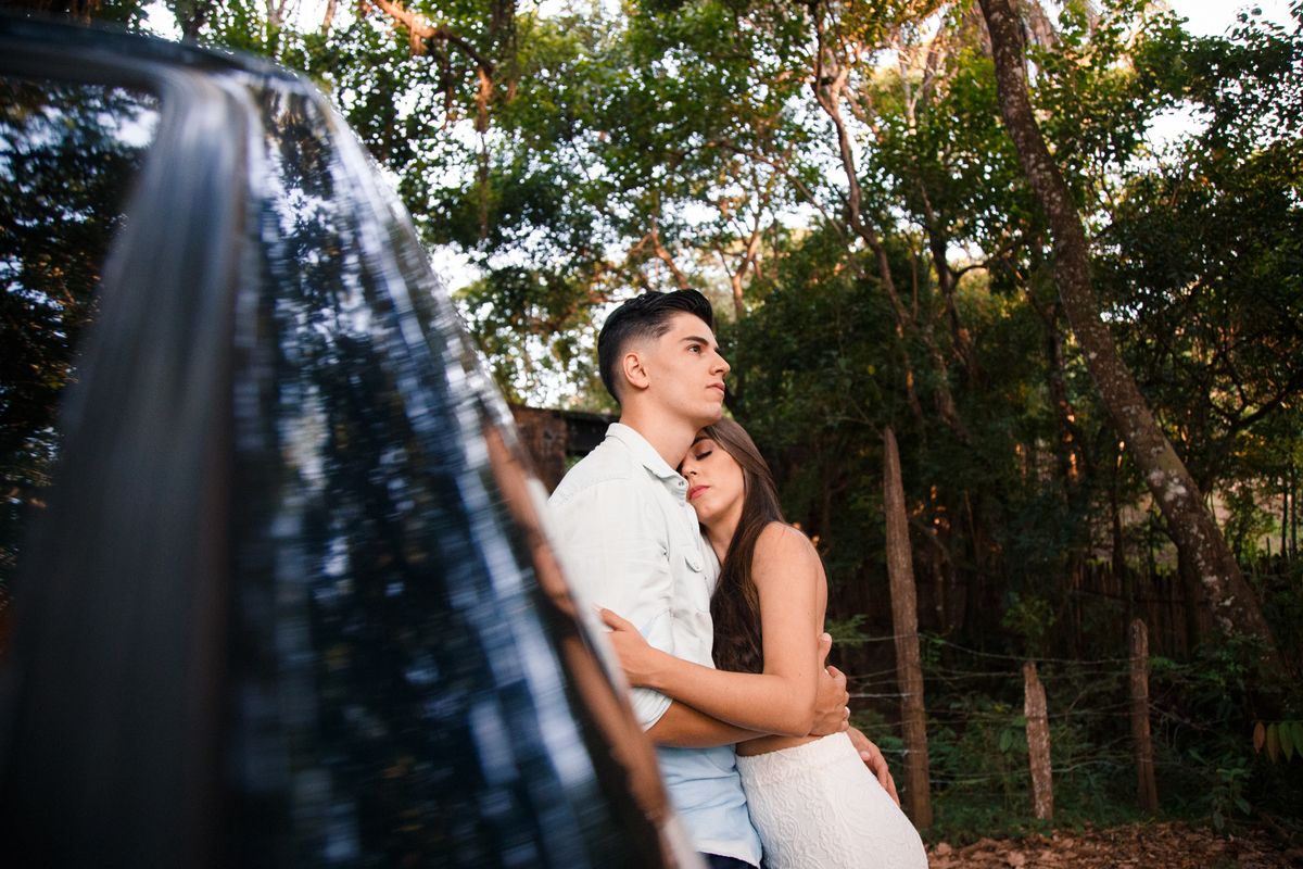 Casal se abraçando, um de frente para o outro, a moça deitada no peito do rapaz que está encostado na porta do carro olhando a paisagem, distraído.