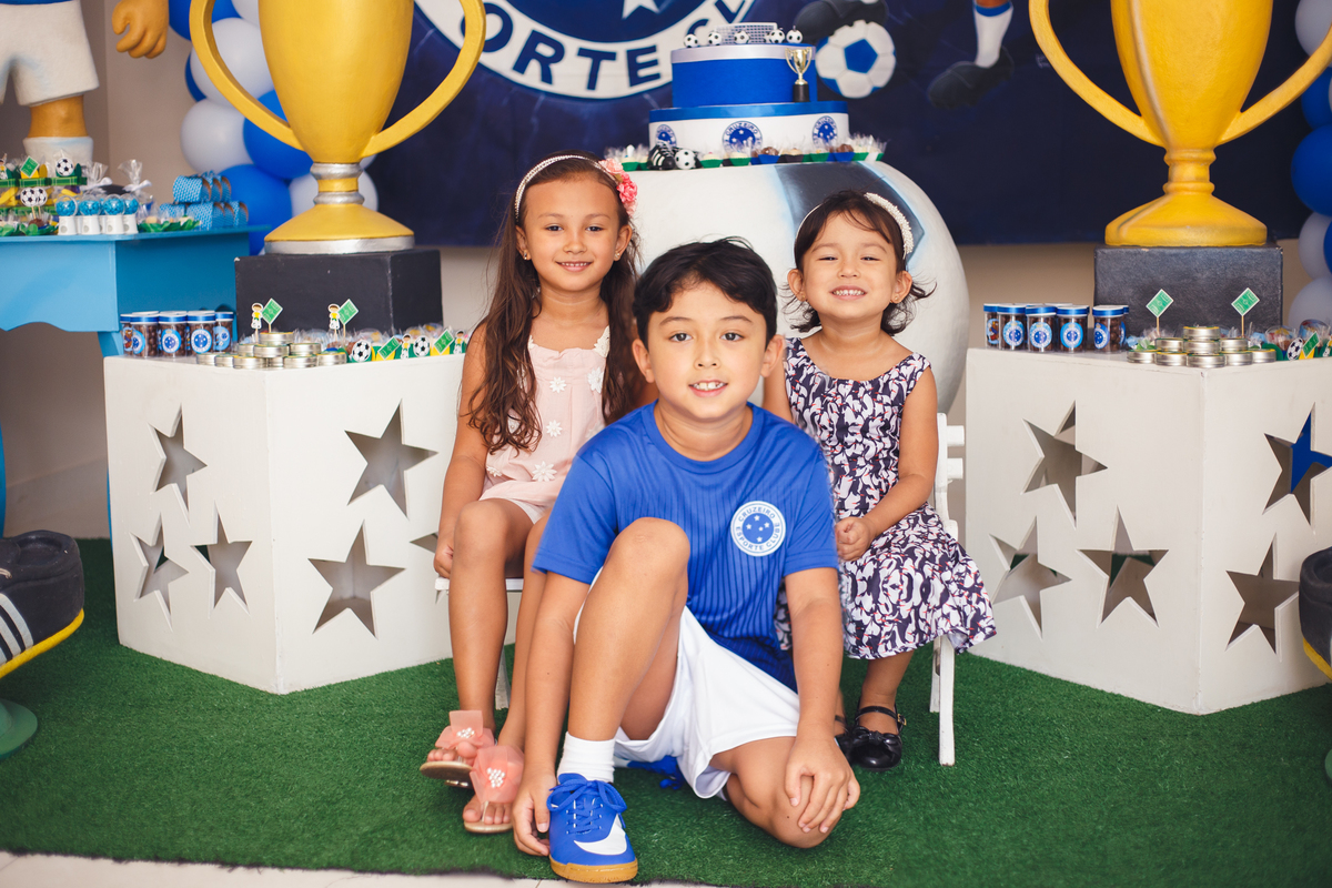 Três irmãos, um menino e duas meninas, posando para foto de aniversário em frente à mesa do bolo.