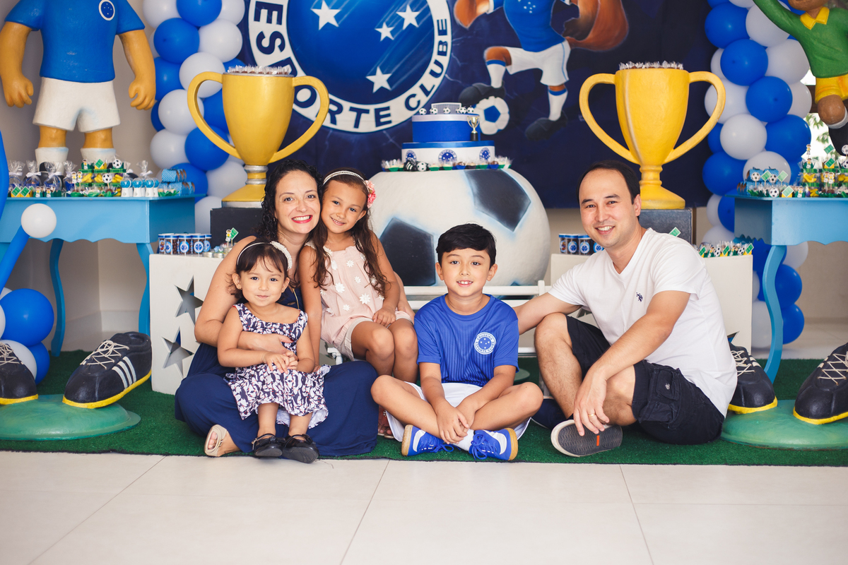 Família reunida na frente da mesa do bolo, todos sentados no chão, pai, mãe e três filhos, um menino e duas meninas.