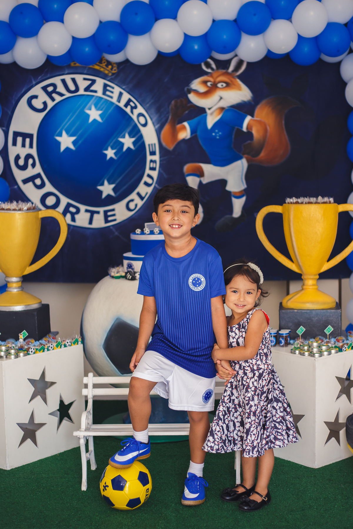 Aniversariante com a irmã mais nova em frente à mesa do bolo, ambos de pé, ele com uma bola e um dos pés em cima da bola, ambos sorrindo.