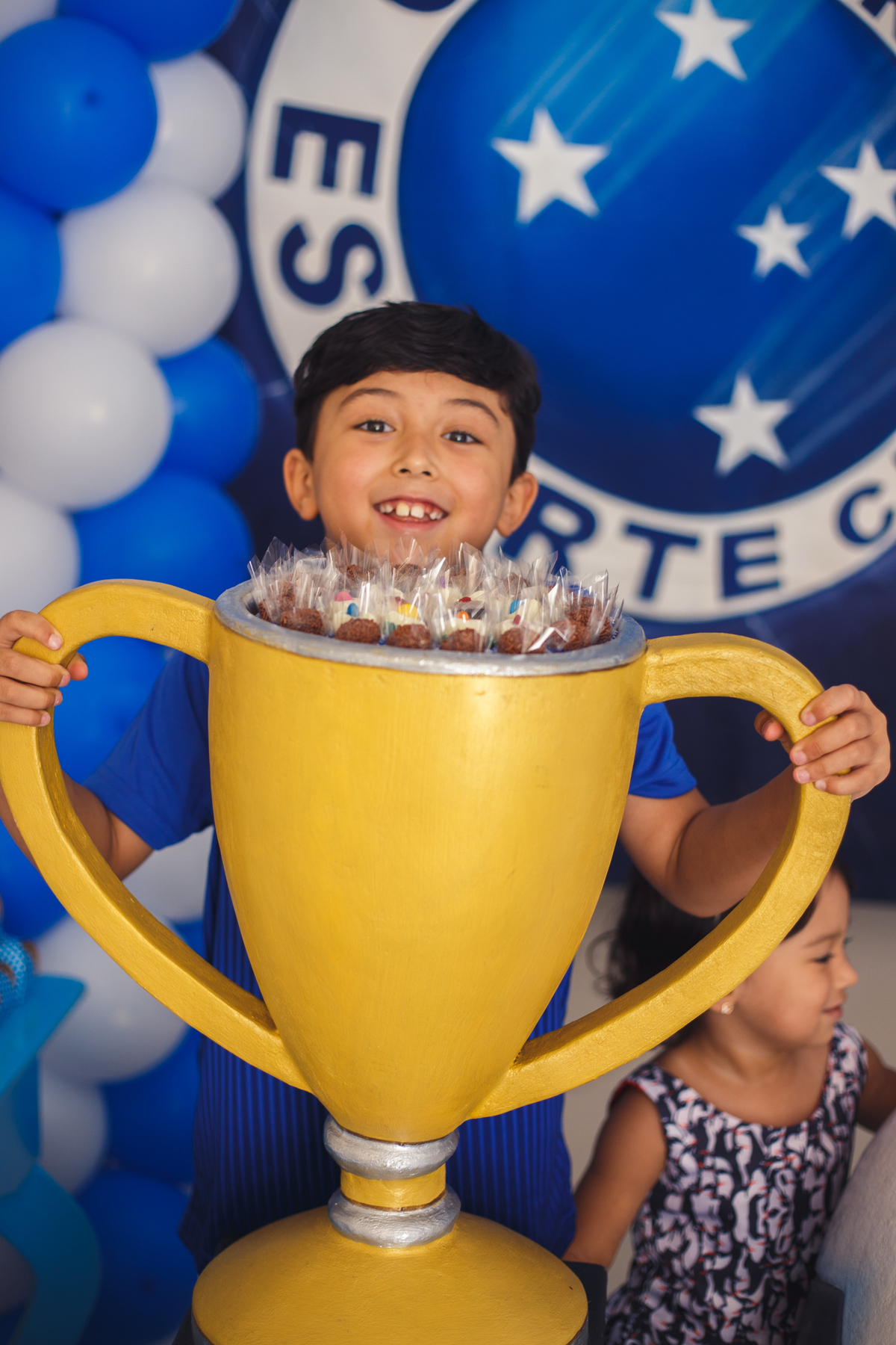 Aniversariante sorrindo atrás de uma taça de campeão, cheia de doces, que enfeita a mesa do bolo, segurando ambas as alças.