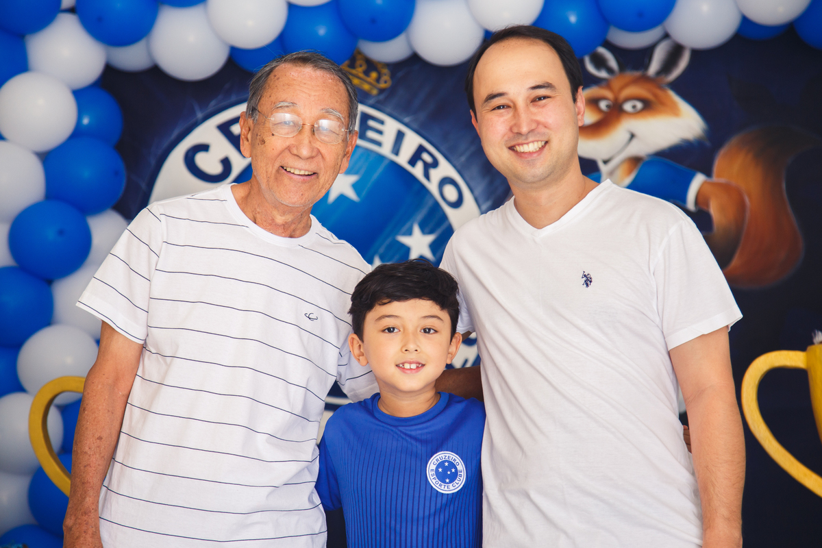 Três gerações, pai, filho e neto, posando em frente à mesa do bolo no aniversário do neto.