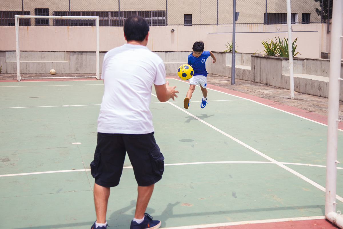Criança ao fundo dando um chute em direção ao gol, onde seu pai está defendendo, de costas, em primeiro plano. 