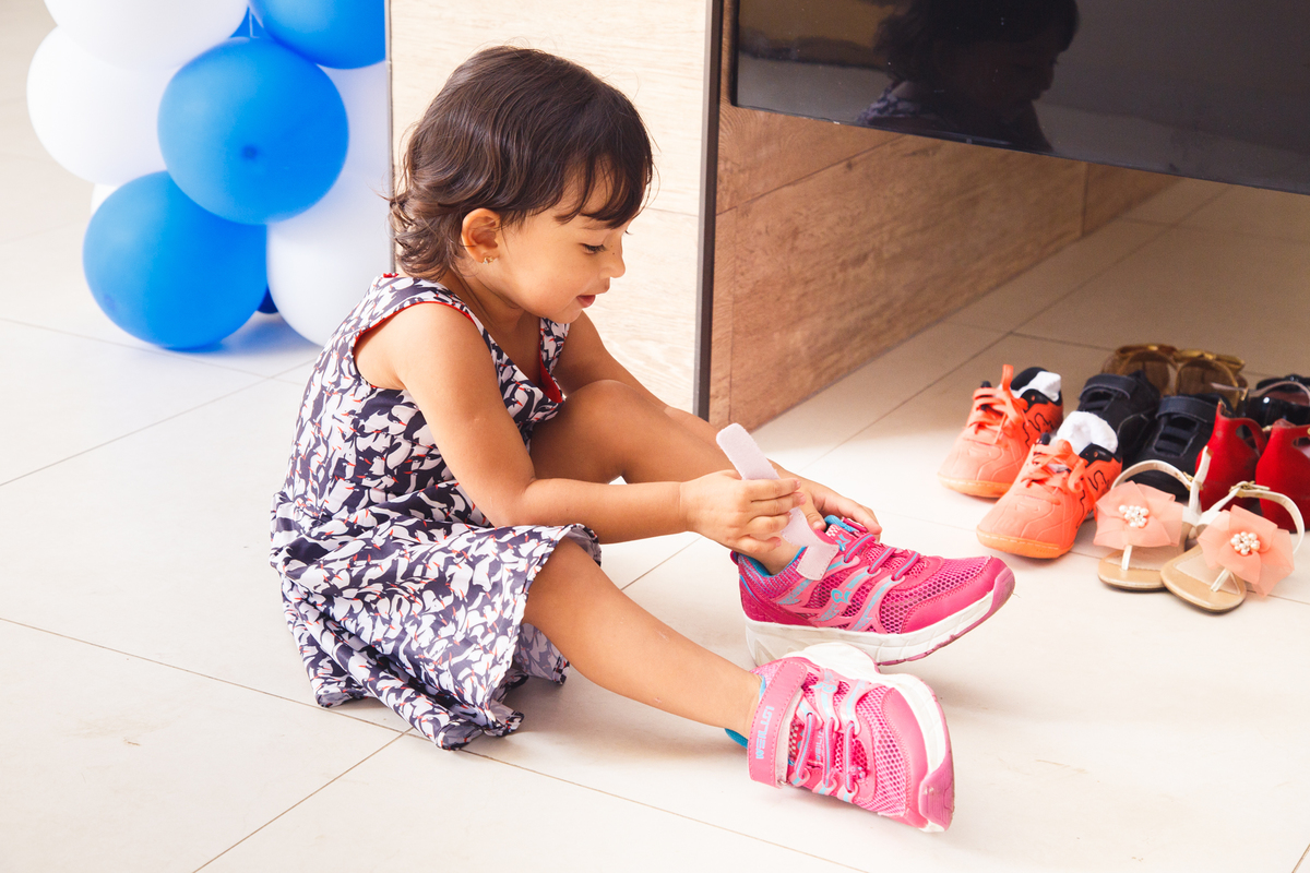 Criança calçando um tênis, muito maior que seu pé, com os pés trocados, o direito no esquerdo e o esquerdo no direito.