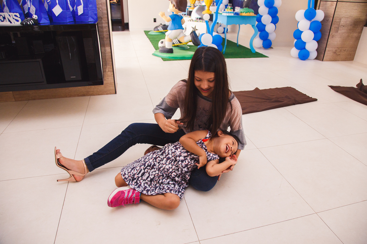 Criança usando um tênis muito maior que seu pé, com rodinhas no calcanhar, deitada no chão, no colo de uma adolescente e gargalhando.
