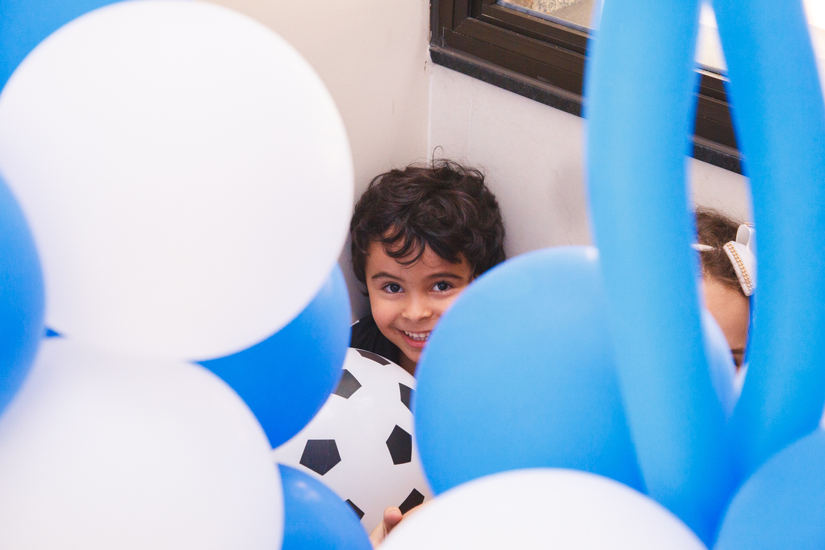 Criança escondida atrás de vários balões, com uma cara sapeca sorrindo e olhando para a foto.