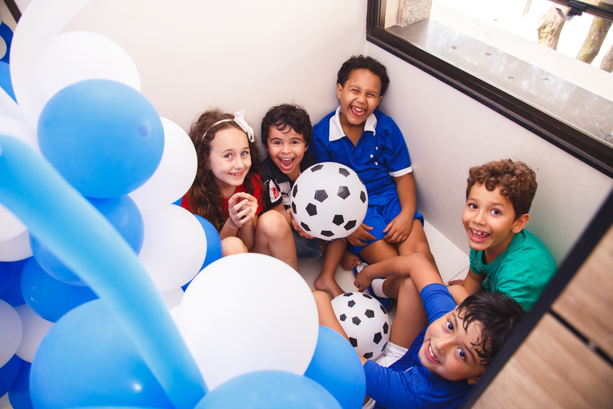 Um grupo de crianças escondido atrás de um arranjo de balões, todos olhando para a foto, fazendo gracinhas e sorrindo.