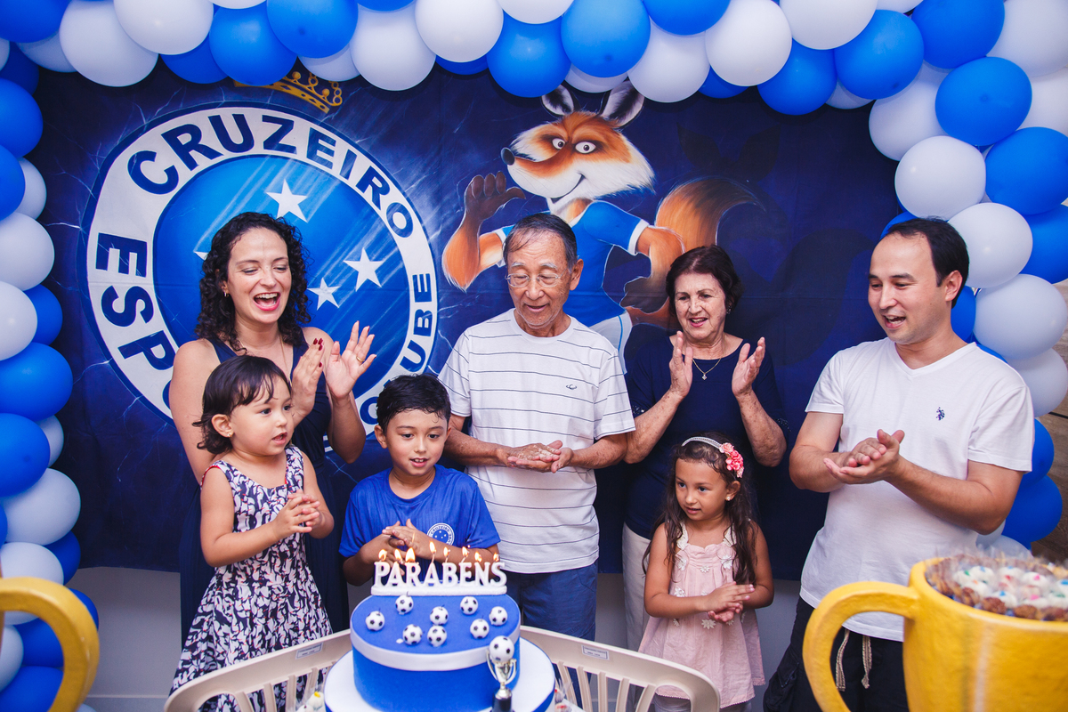 Família reunida atrás da mesa do bolo cantando parabéns para o avô do aniversariante.