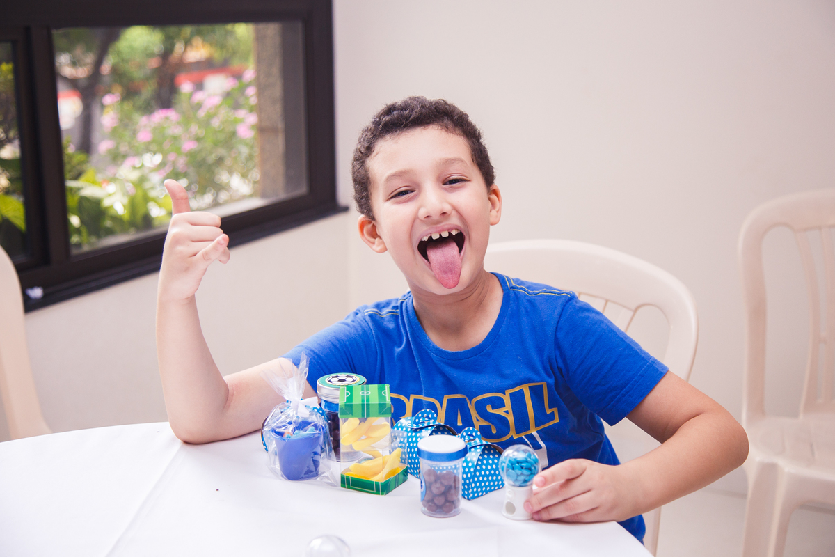 Criança em uma festa de aniversário infantil sentado com vários potinhos de bala em sua frente fazendo uma careta com a língua para fora e um joia com o polegar olhando para a fotógrafa.