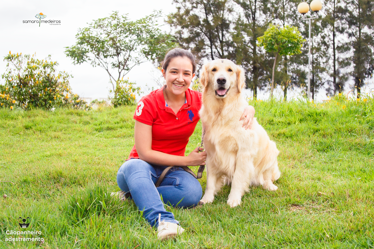 Cachorro golden sentada na grama com sua tutora, se abraçando ambas olhando para a câmera, com árvores ao fundo, em parque de Belo Horizonte.
