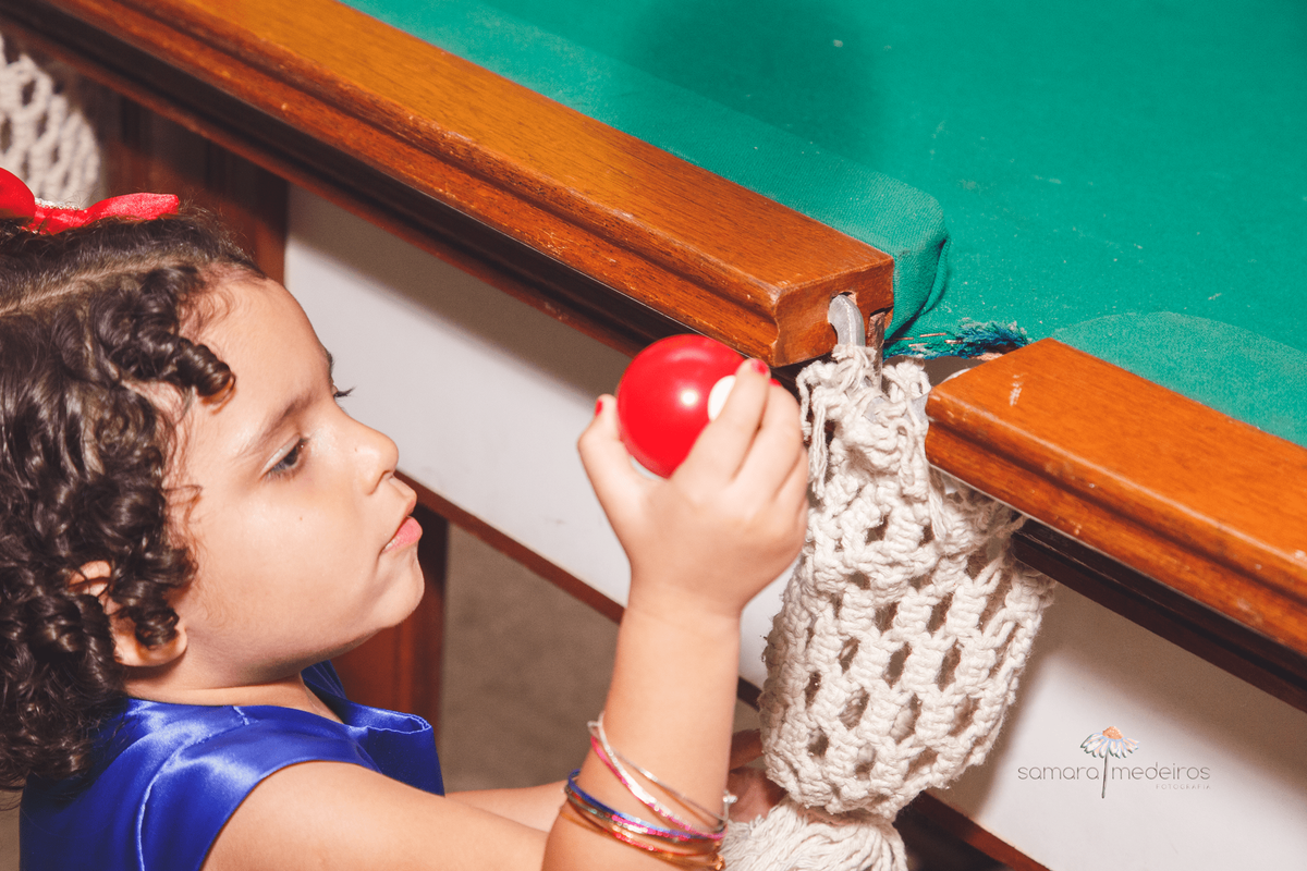 Criança com roupa de branca de neve pegando uma bola de sinuca vermelha e olhando para ela.