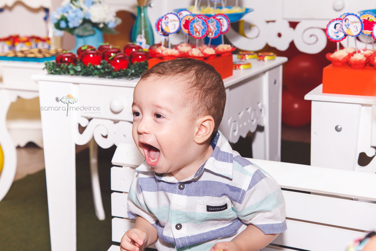 Bebê sentado no banquinho na frente da mesa do bolo, dando uma enorme gargalhada.
