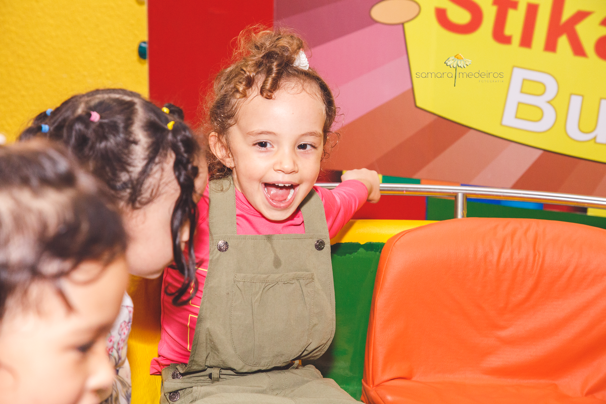 Criança sentada no gira gira, segurando na barra de apoio, olhando para outra criança e com a boca aberta sorrindo.