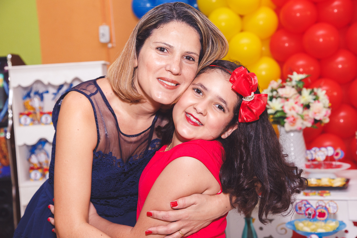Mãe abraçando sua filha durante aniversário infantil, com a mesa do bolo ao fundo.
