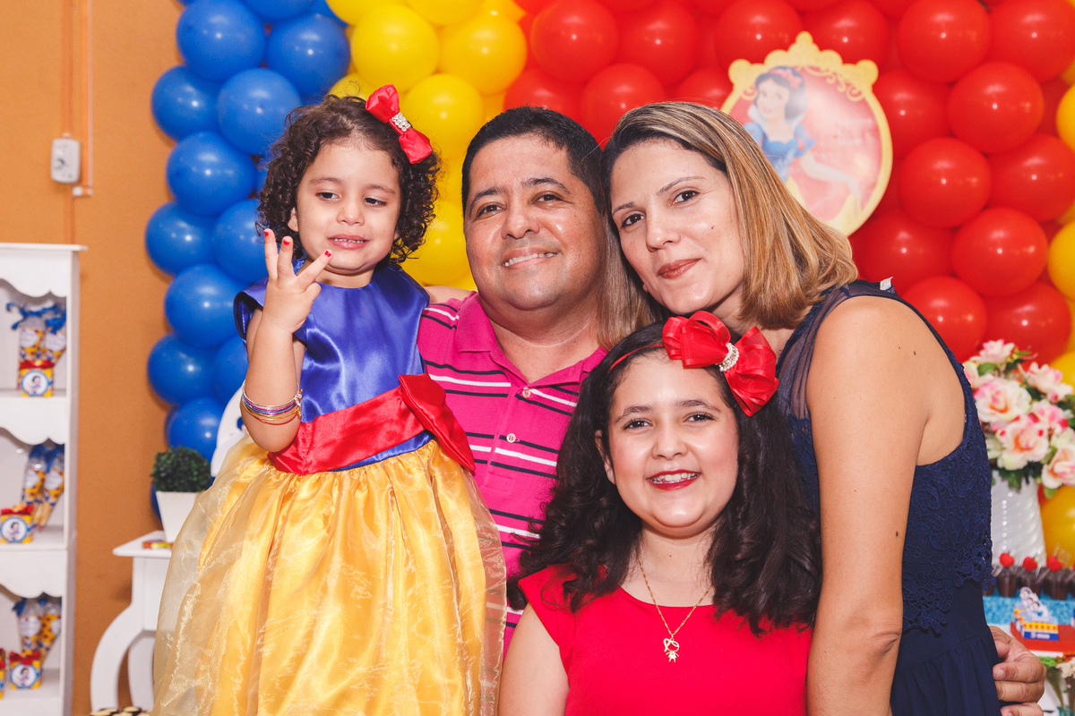 Família reunida, pai, mãe e duas filhas, em frente à mesa do bolo no aniversário infantil das meninas.