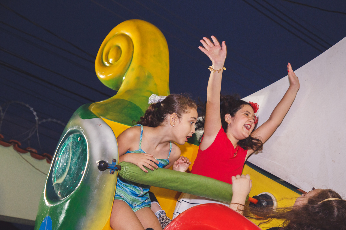 Duas crianças no brinquedo estilo barco que vai e vem, uma delas com os braços levantados e gritando, sorrindo, a outra com cara de susto.