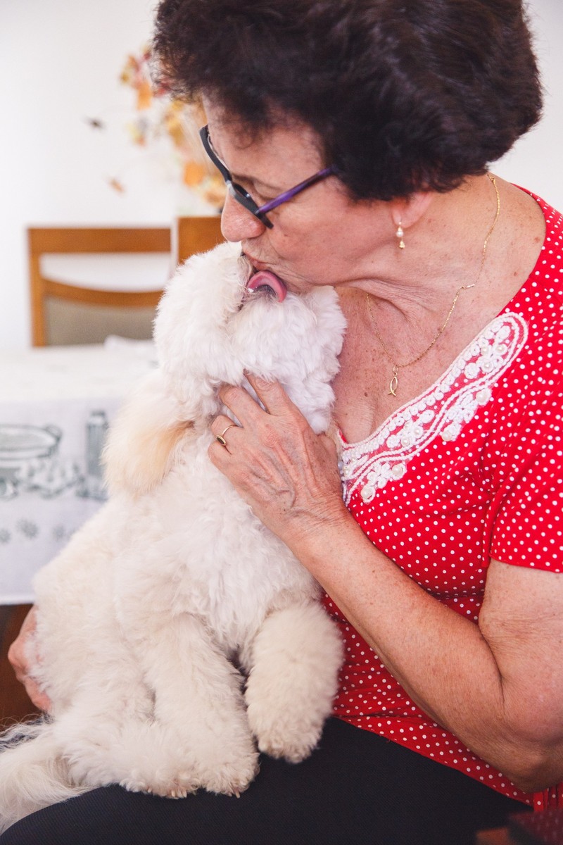 Tutora sentada em uma cadeira com sua cachorrinha poodle no colo, ganhando uma lambida no rosto.