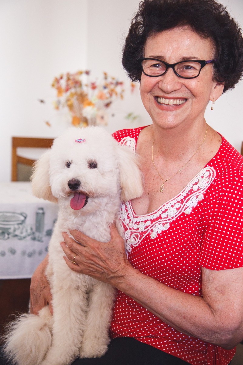 Tutora e com sua cachorrinha poodle no colo, olhando para a câmera e sorrindo, sentada em uma cadeira em sua sala.