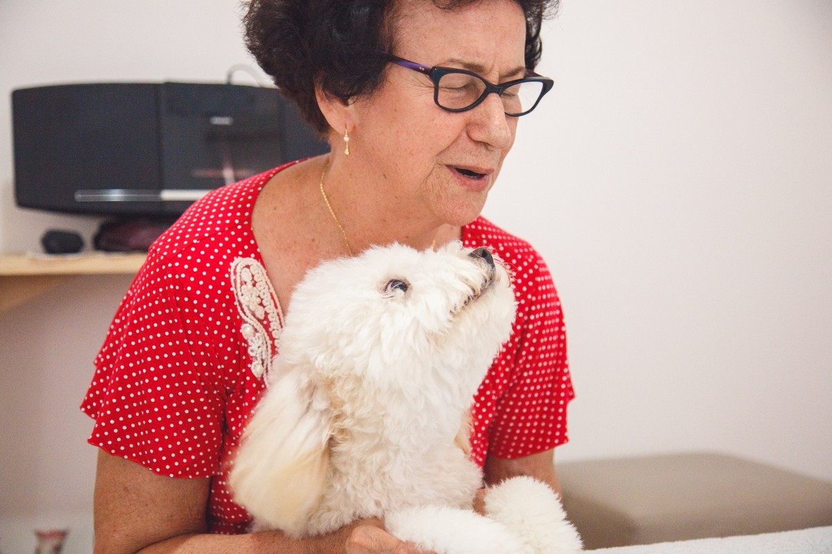 Tutora de uma pet ajoelhada na beira da cama fazendo uma oração com a poodle entre seus braços, apoiada na cama e olhando para a tutora.