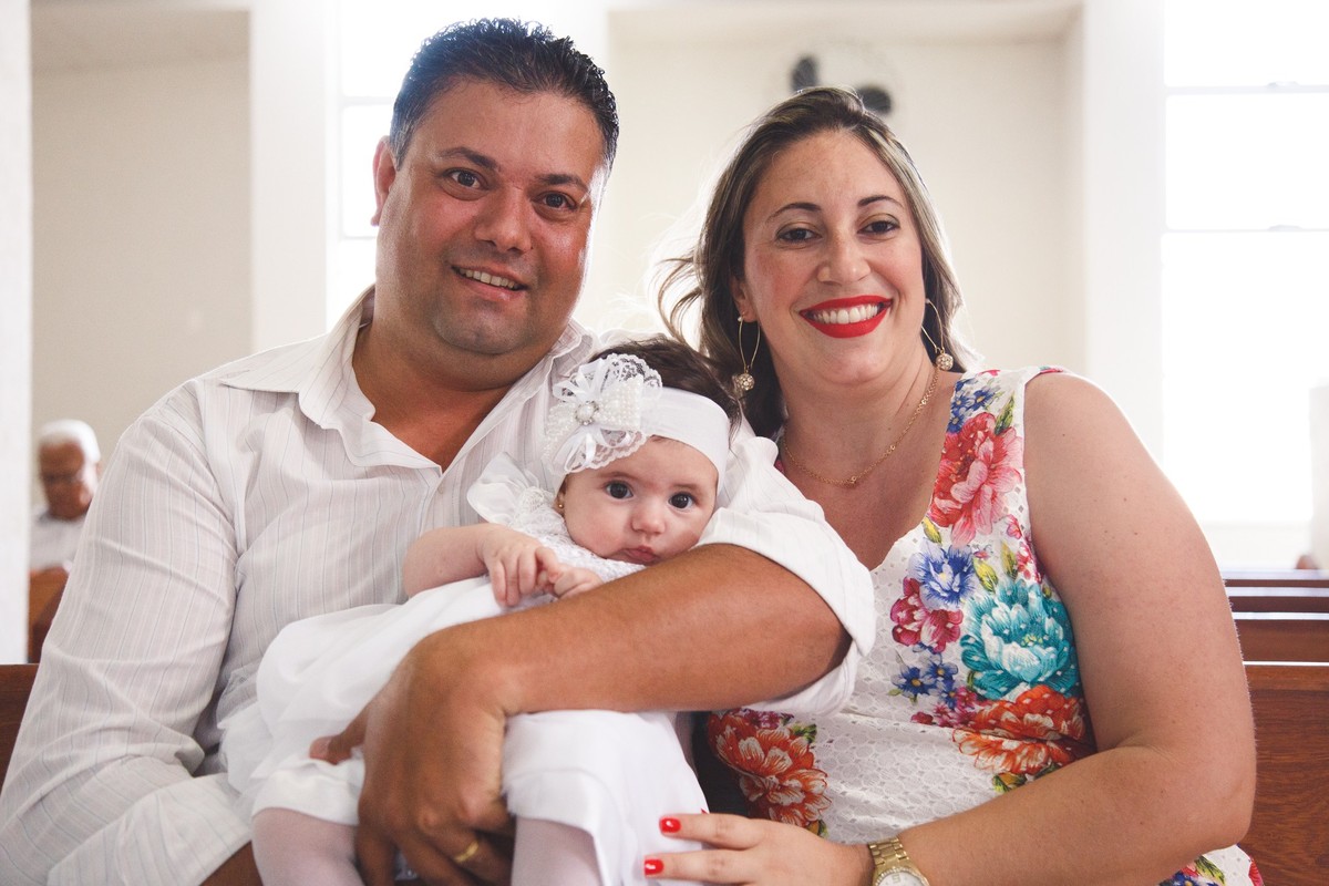 Pai e mãe com a bebê sentados na igreja aguardando o início do batizado da filha, olhando para a câmera e sorrindo.