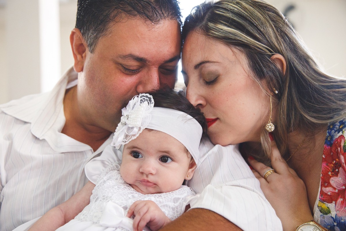Pai e mãe cheirando a cabeça da bebê antes do batizado, enquanto estão sentados no banco da igreja.