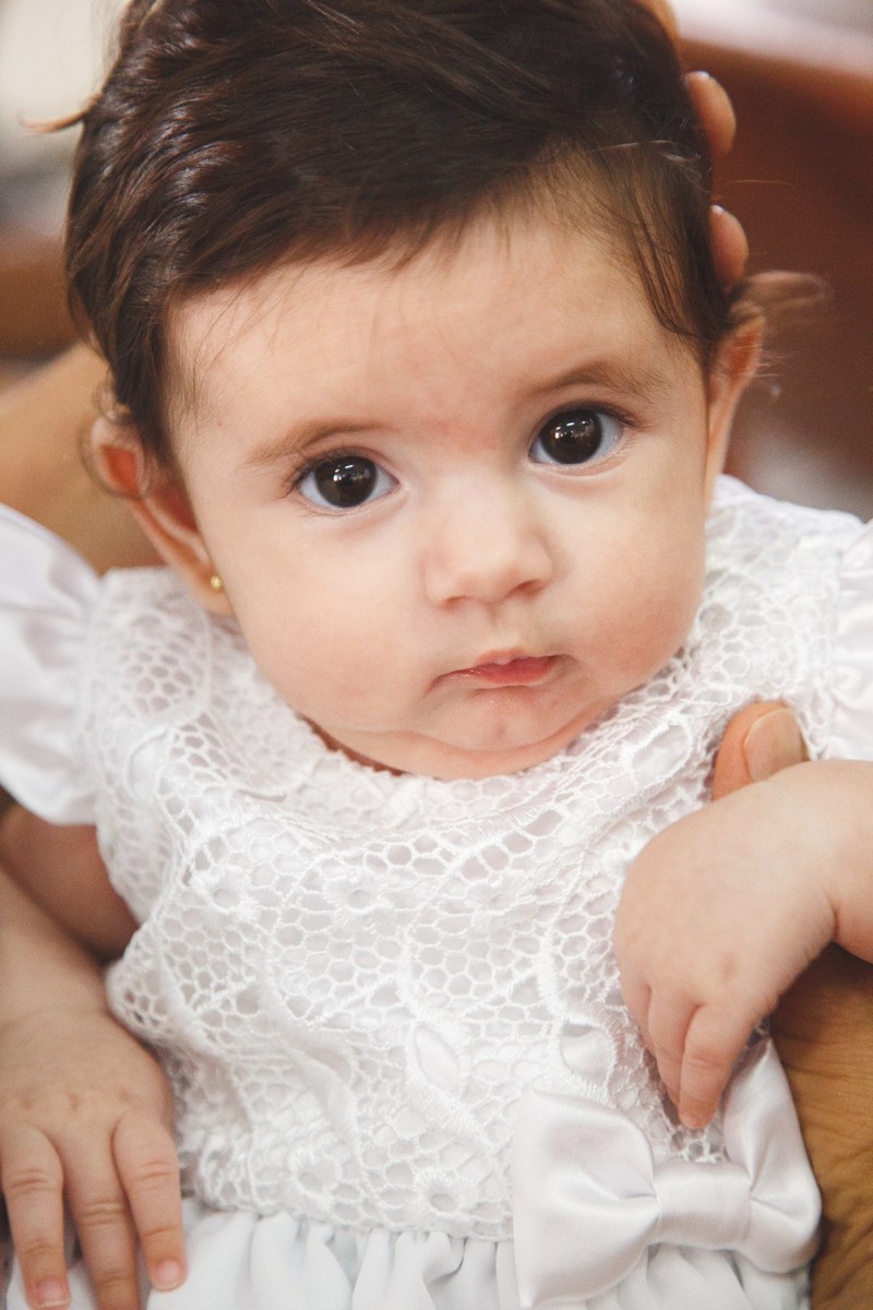 Close no rosto da bebê, que observa a foto, durante sua cerimônia de batizado.
