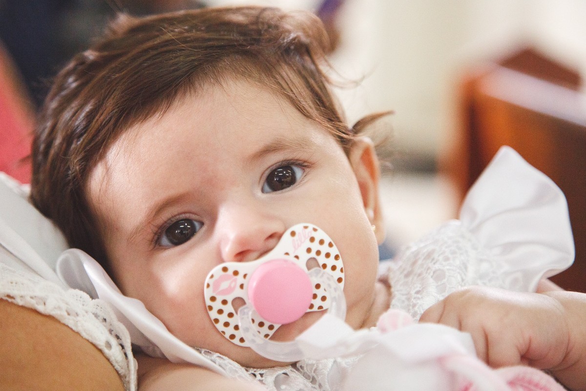 Bebê deitada no colo de sua avó, usando um bico e olhando para a foto, durante seu batizado.