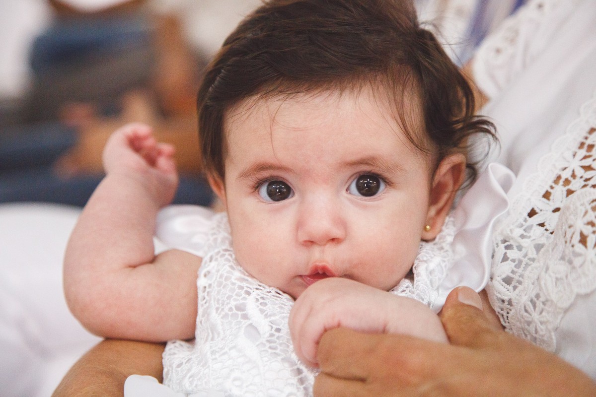 Close na bebê, no colo de sua avó, fazendo uma careta bonitinha após sua cerimônia de batizado.