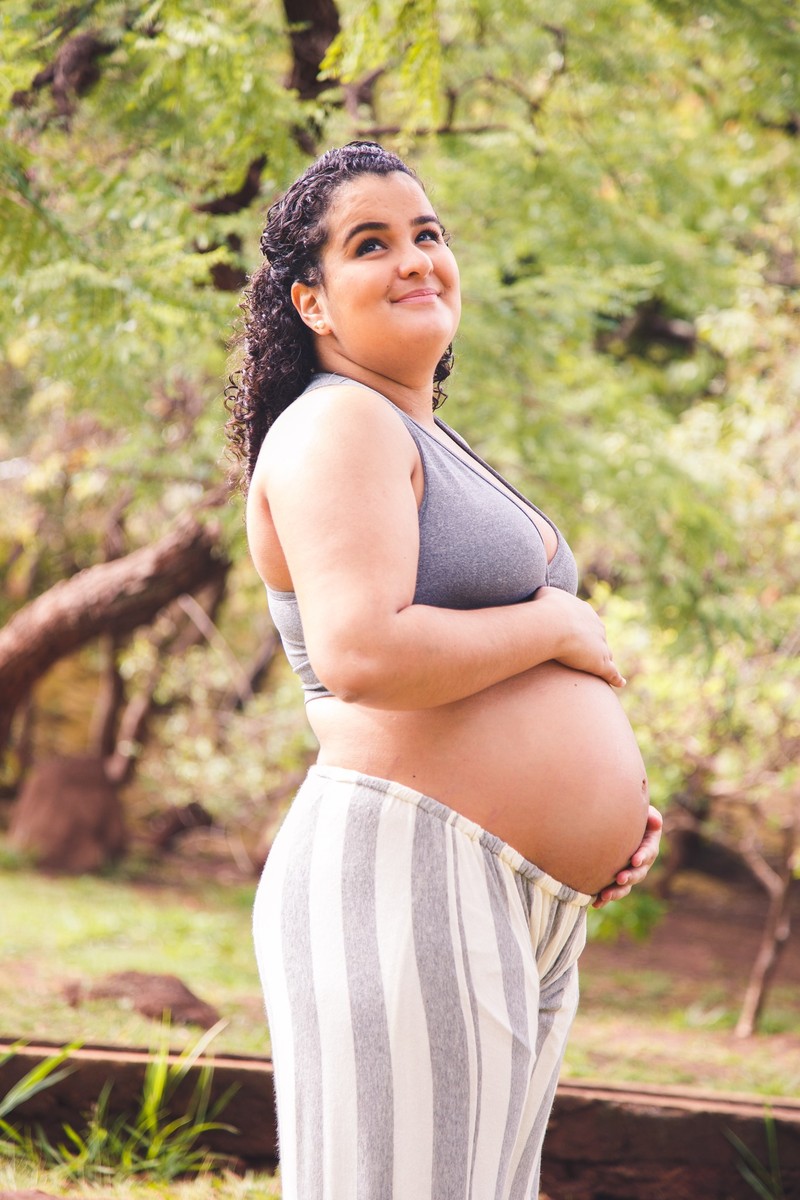 Gestante de lado, com as mãos apoiadas na barriga, fazendo carinho e com uma expressão pensativa.