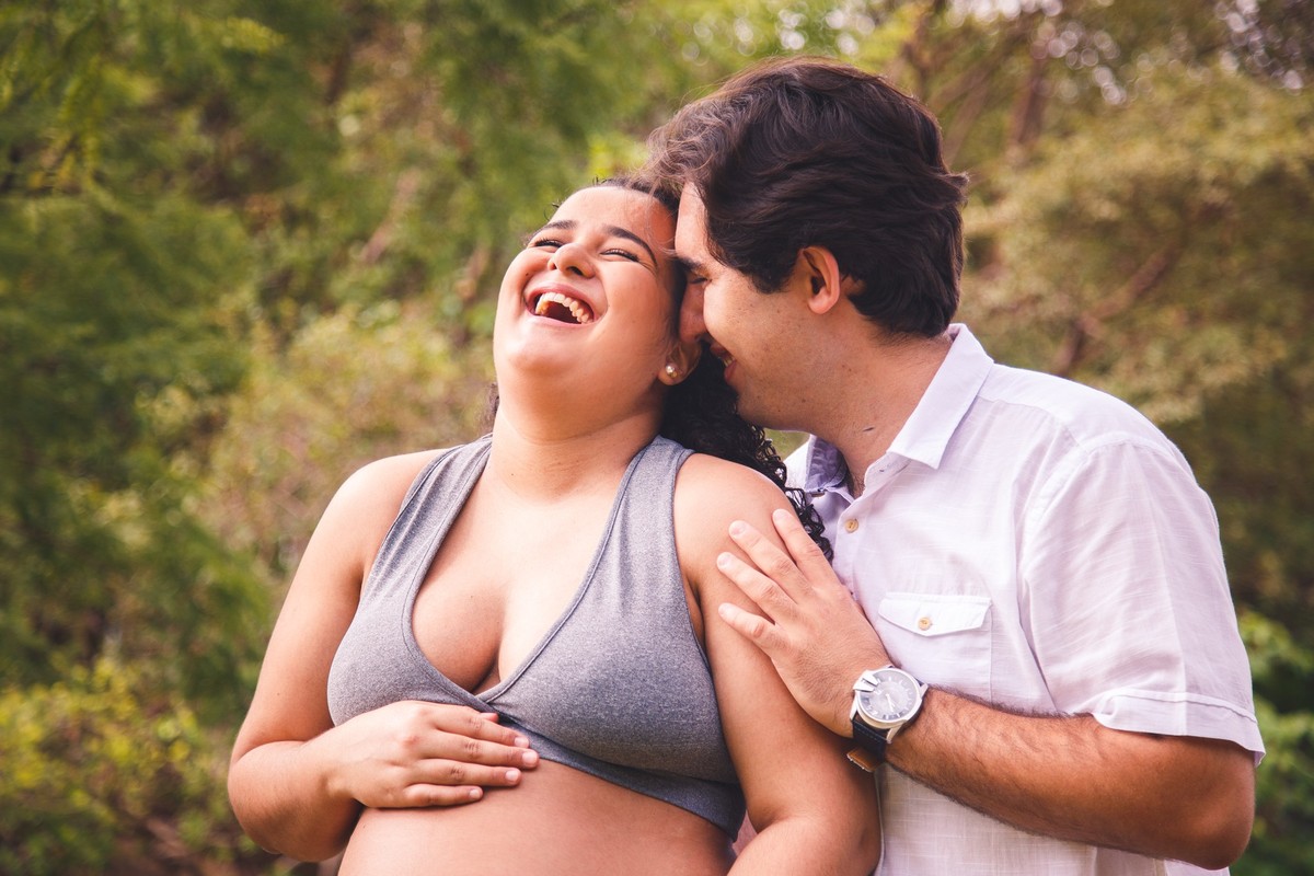 Casal de pé abraçados, o marido falando no ouvido da gestante e ela gargalhando.