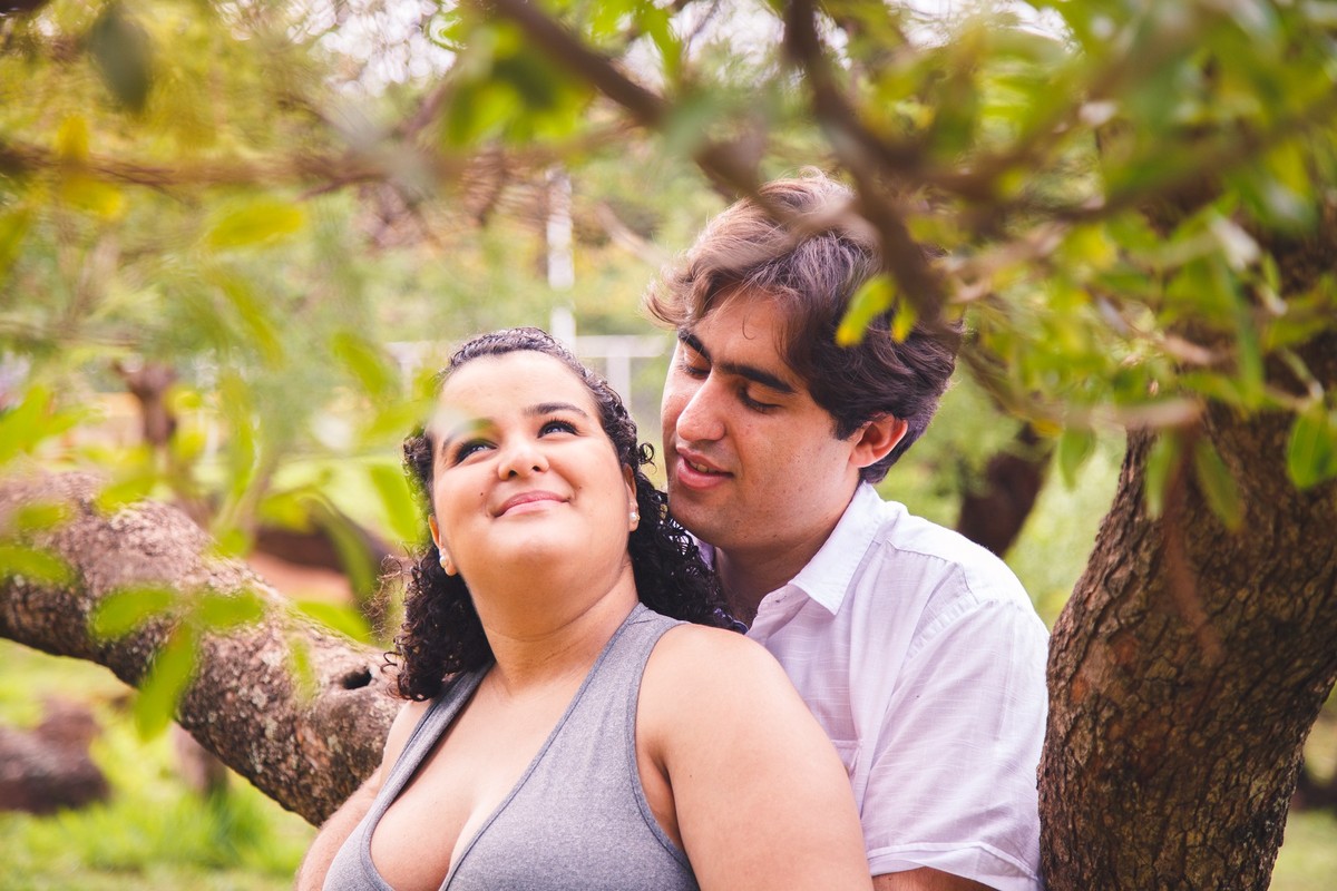 Casal em ensaio gestante, apoiados em uma árvore e um apoiado contra o outro, o marido olhando para a gestante com expressão feliz.