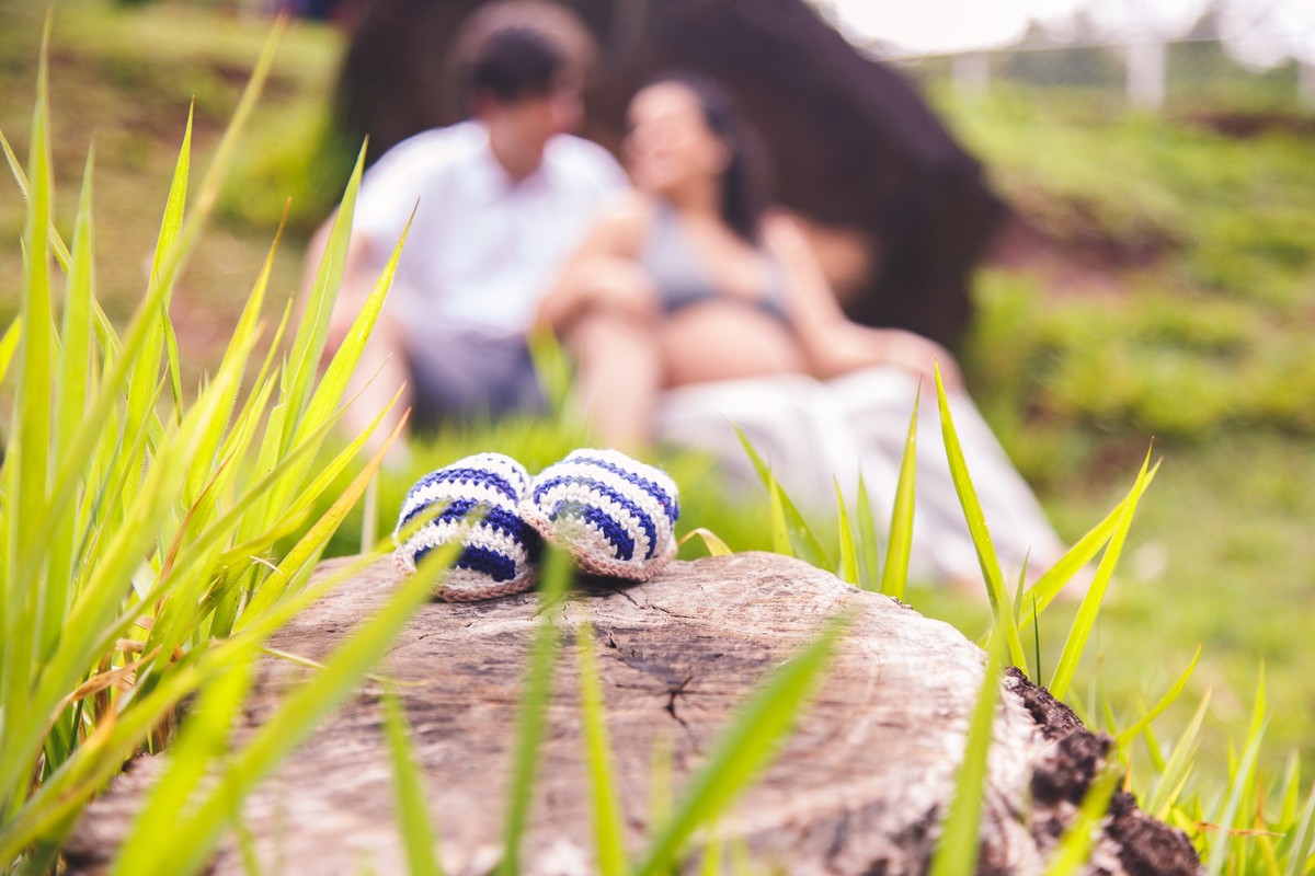 Sapatinho de bebê apoiado em uma pedra em primeiro plano e o casal no fundo desfocado.