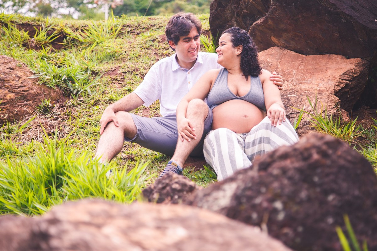 Casal sentado na grama, se olhando, a gestante apoiada no marido, ambos sorrindo.