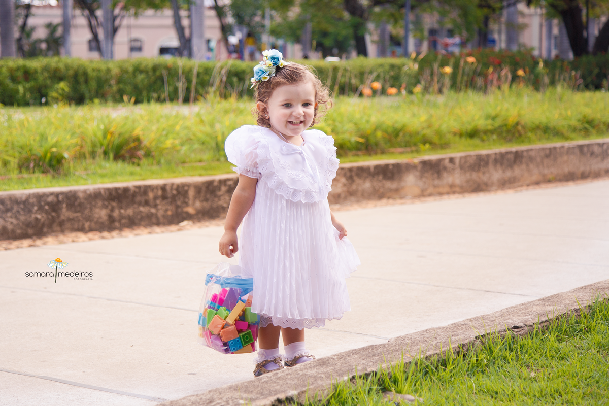 Criança parada de pé em uma praça de Belo Horizonte, sorrindo e segurando uma bolsinha de brinquedo.