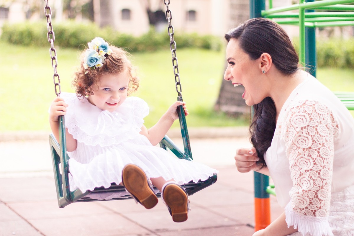 Menina de 2 anos brincando no balanço, em uma praça de BH, enquanto sua mãe está a seu lado sorrindo e brincando.
