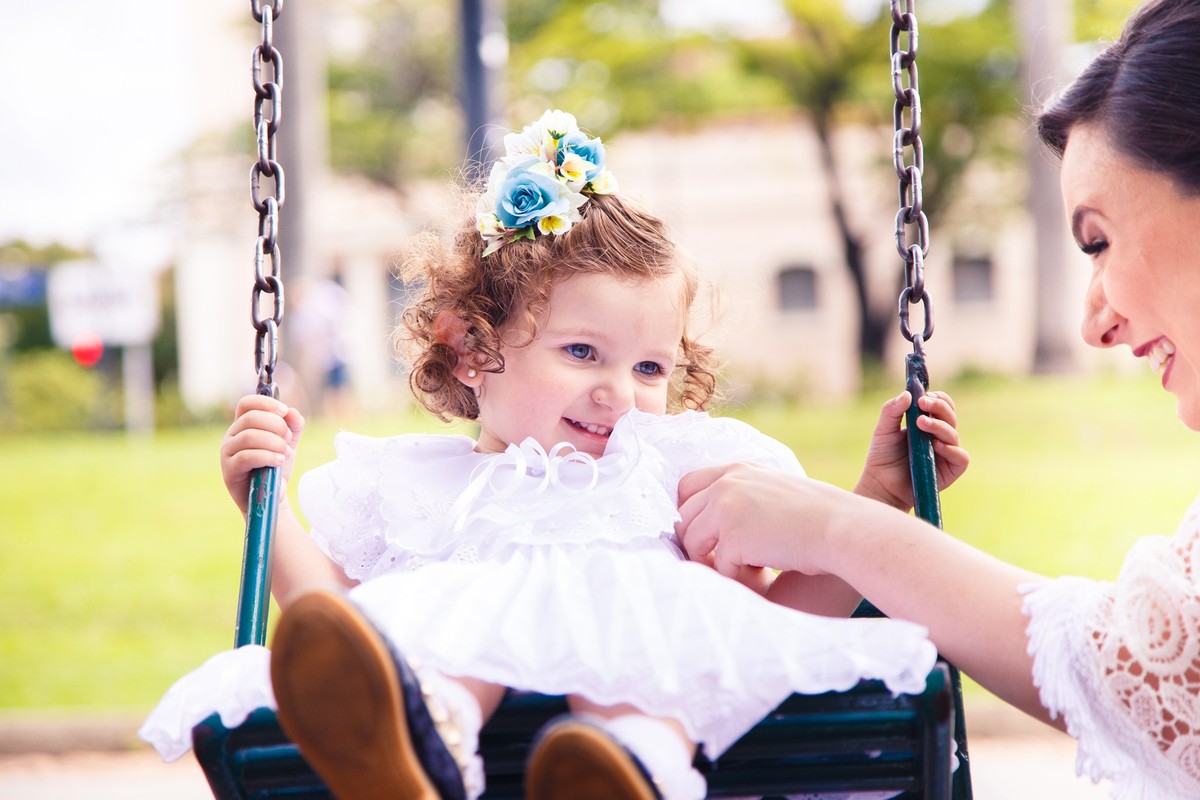 Mãe fazendo cócegas na sua filha, que está brincando em um balanço de parquinho infantil, ambas sorrindo.