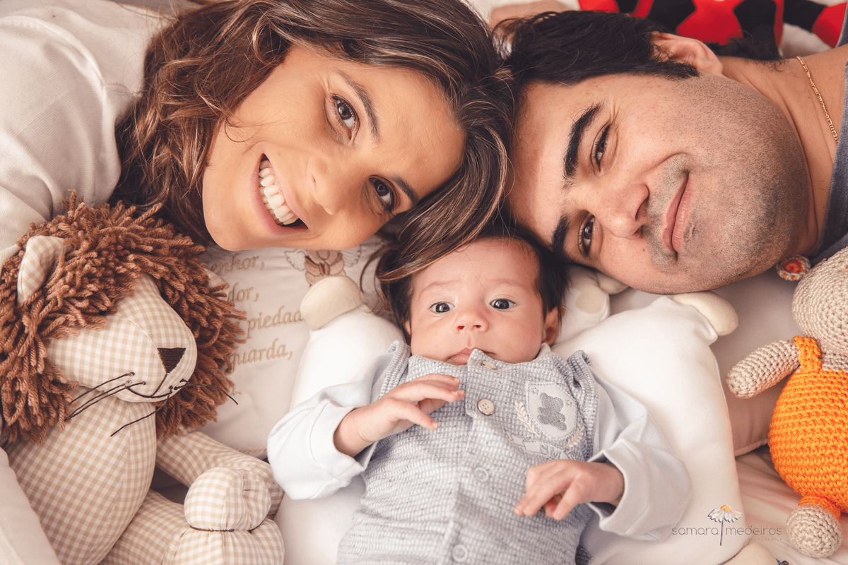 Mãe e pai ao lado de seu filho recém nascido, os três com o rosto próximo uns dos outros, deitados na cama.