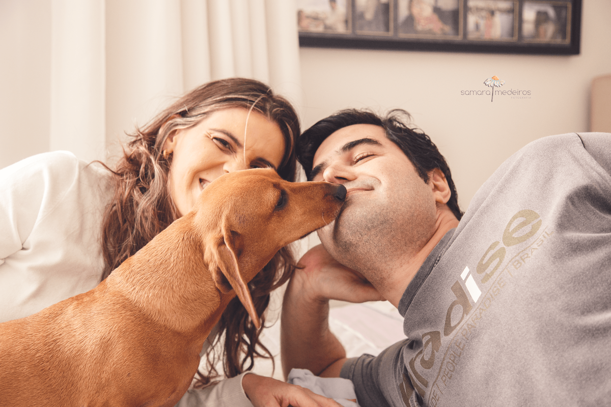 Pai e mãe deitados na cama, o pai recebendo uma lambida no rosto da cachorrinha dachshund da família.